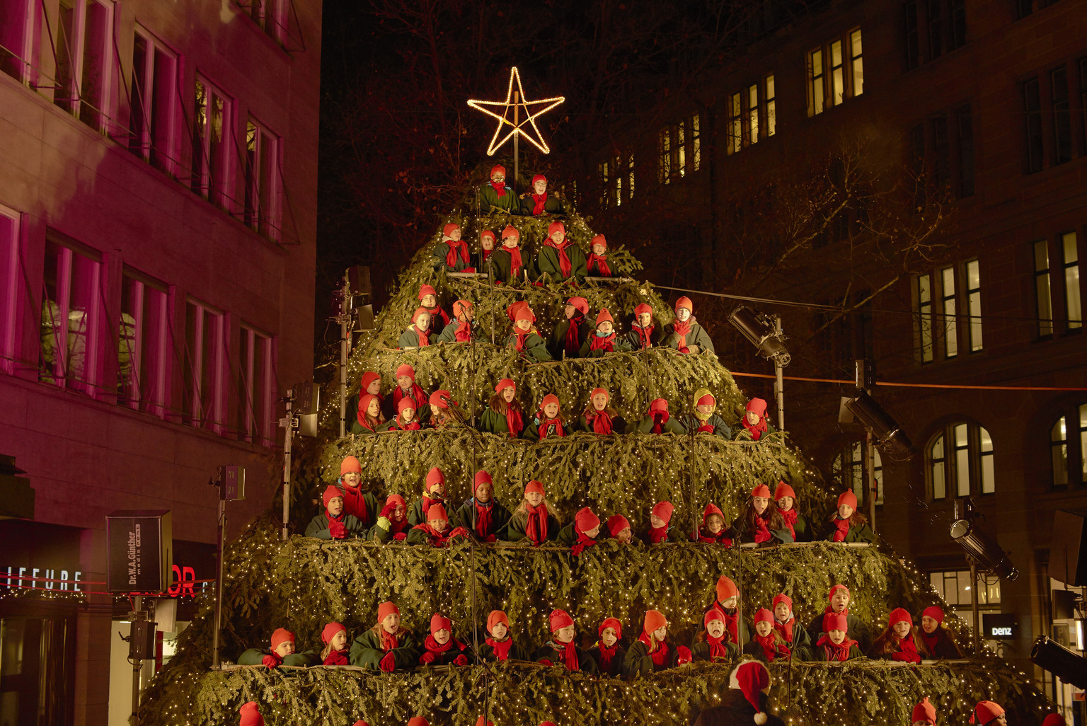 Der Singing Christmas Tree auf dem Werdmühleplatz: ein baumförmiges Gerüst, auf dessen Stufen ein Chor in roten Mützen singt